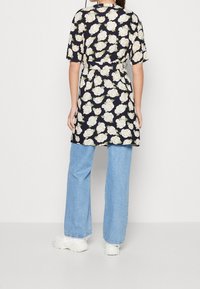 Květinové šaty s krátkým rukávem a páskem v pase na osobě, umístěné na bílém pozadí. Šaty mají černý základ s bílým a zeleným květinovým vzorem, kombinované se světle modrými širokými džíny a bílými teniskami.