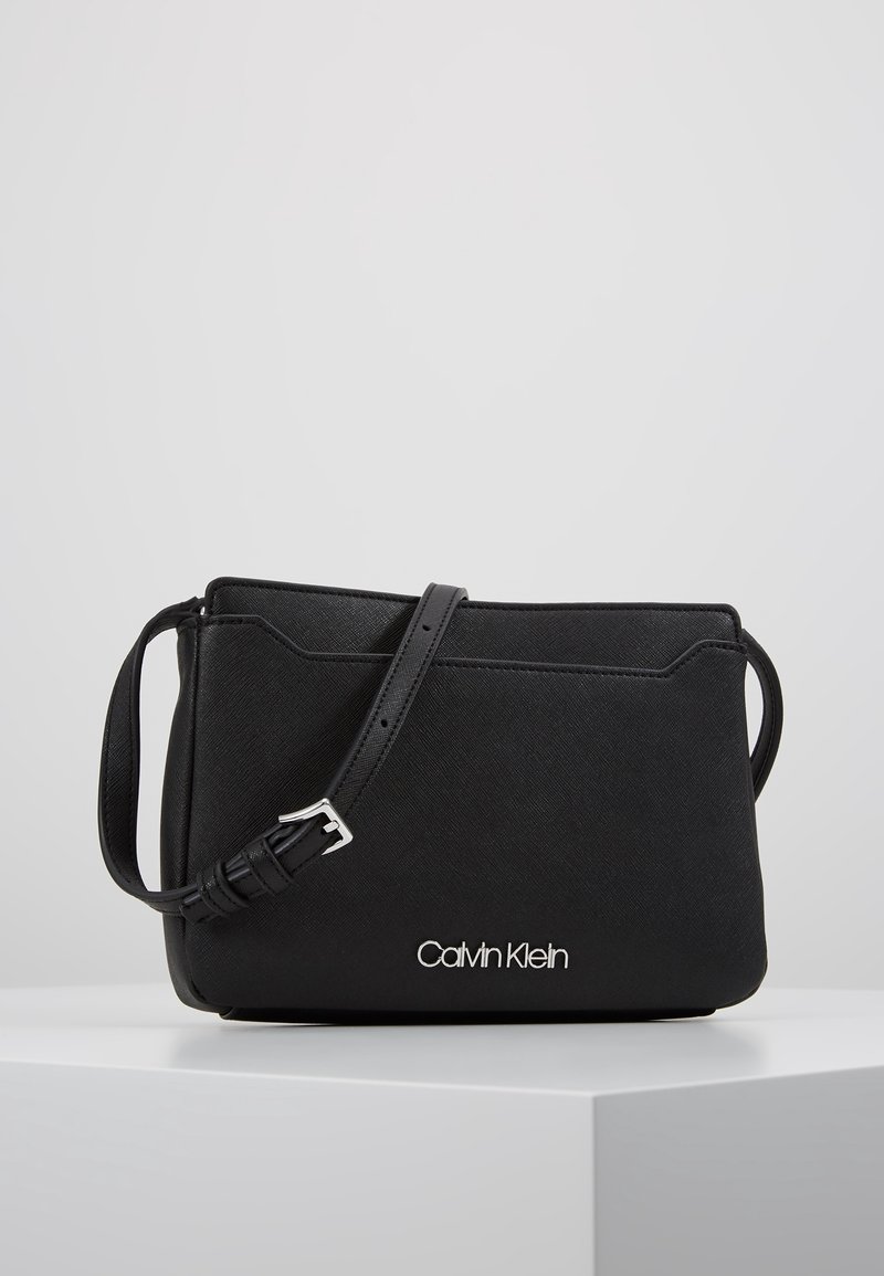Musta Calvin Klein olkalaukku säädettävällä hihnalla ja hopeisella logolla, esillä valkoisella jalustalla harmaan taustan edessä.