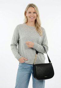 Vrouw met blond haar, gekleed in een grijze trui en blauwe spijkerbroek, met een zwarte leren schoudertas met gouden kettinkdetail.