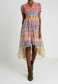 Robe colorée à motifs avec des manches courtes bouffantes, un décolleté en V et un ourlet asymétrique. Présente des motifs géométriques dans des teintes de rose, bleu et jaune.