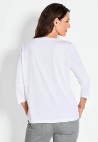 Weißes Langarm-T-Shirt mit lockerer Passform, Rundhalsausschnitt und strukturiertem Stoff, getragen mit grauen Hosen, von hinten gesehen.