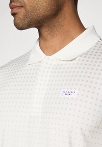 Balta polo krekla ar smalku punktiņu rakstu, klasisku apkakli un zīmola uzlīmi uz krūtīm. Izgatavots no viegla materiāla.