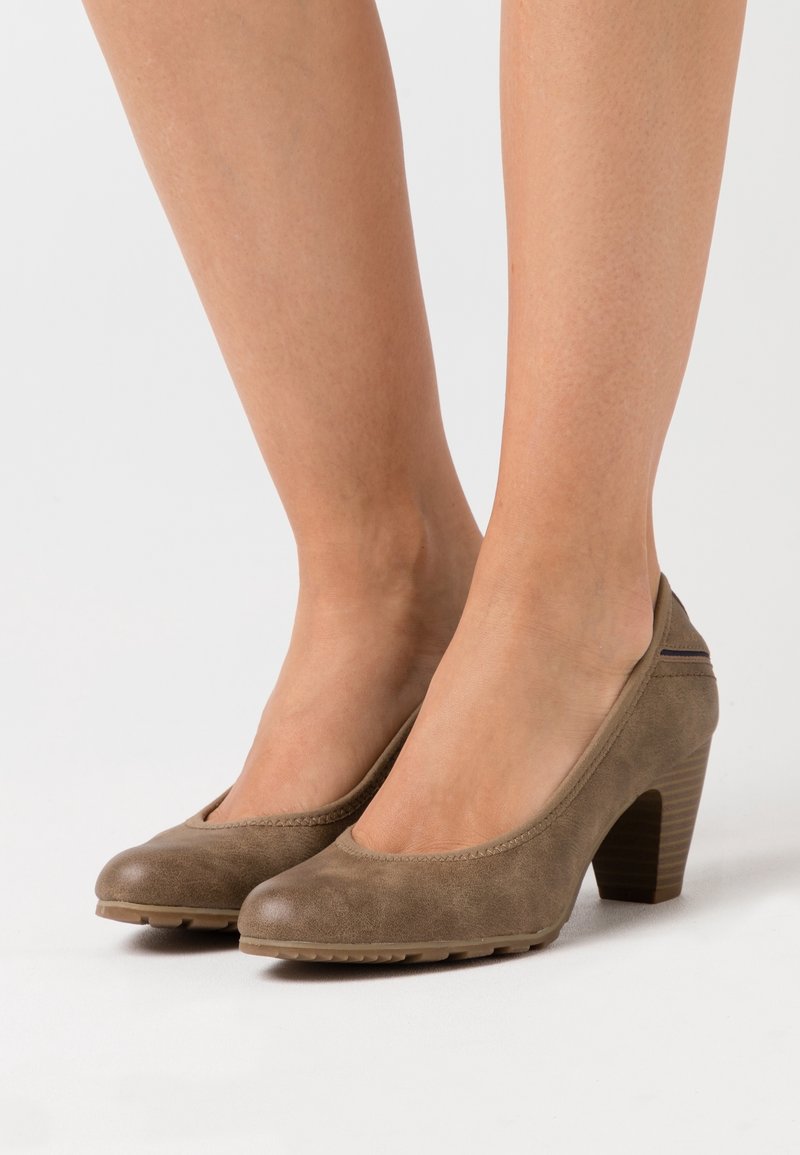 Jambes de femme portant des escarpins taupe en suède à bout rond et talon moyen avec semelles en caoutchouc, debout sur une surface blanche.