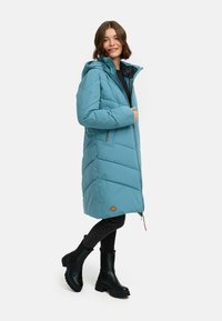 Jasnoniebieska ocieplana kurtka z kapturem, z suwakiem z przodu, bocznymi kieszeniami i pikowanym wzorem. Połączona z czarnymi spodniami i butami.