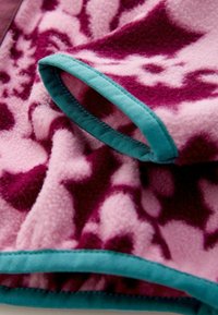 Fleece-Stoff in Pink mit tiefviolettem abstraktem Muster; eingefasst mit einem türkisblauen Rand. Weiche Textur mit glatter Oberfläche in schichtweisen Falten.