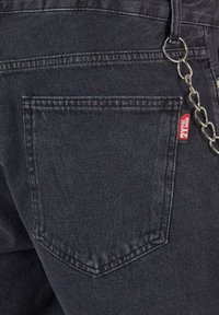 Jeans de mezclilla negra con un bolsillo trasero visible, accesorio de cadena y una pequeña etiqueta roja. Tejido suave y texturizado con un diseño robusto.