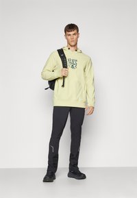 Hanorac cu glugă de culoare galben deschis, cu un logo, asortat cu pantaloni negri, strâmți, ce au accente reflectorizante și pantofi sport negri.