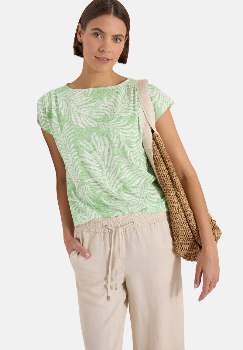 Jeune femme portant un haut vert et blanc à motif de feuilles, un pantalon beige à cordon, et un sac à bandoulière tressé marron.