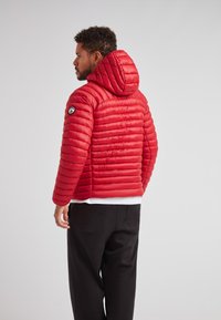 Giacca imbottita rossa con cappuccio, caratterizzata da un design trapuntato, finitura testurizzata e logo circolare sulla manica sinistra. Abbinata a pantaloni scuri.