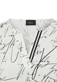 Blouse blanche avec imprimé abstrait en script noir, décolleté en V, et un détail rayé noir et blanc au niveau de l'encolure.