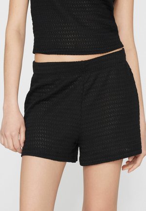 Mujer con pantalones cortos de punto texturizados negros y top corto sin mangas a juego, mostrada desde la mitad del muslo hasta el torso medio.