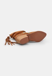 Bota de ante marrón hasta el tobillo con una suela flexible, acentos decorativos en azul y dorado, parte superior fruncida y borlas en los laterales para añadir textura.