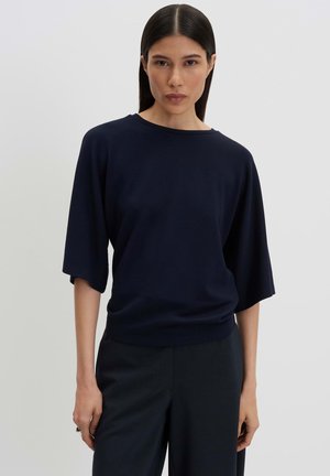 Femme aux longs cheveux foncés portant un haut bleu marine ample à manches aux coudes et un pantalon sombre sur un fond clair uni.