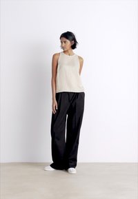 Beige sleeveless top paired with high-waisted black pleated pants and white shoes. Smooth textures and relaxed fit in neutral colors.