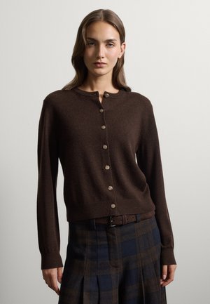 Frau trägt eine dunkelbraune, geknöpfte Strickjacke und einen plissierten karierten Rock mit braunem Gürtel und steht vor einem einfarbigen Hintergrund.