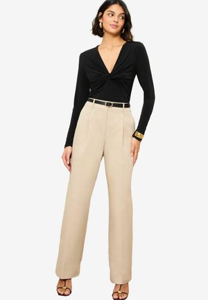 Top noir à manches longues avec un design avant tordu, associé à un pantalon beige taille haute et des sandales à talons noires avec des accents dorés.