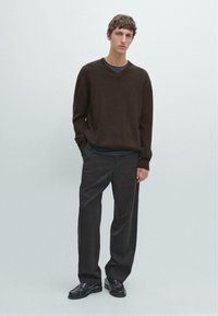 Maglione marrone a maglia con scollo a V, indossato sopra una camicia scura; abbinato a pantaloni scuri larghi e scarpe nere lucide.