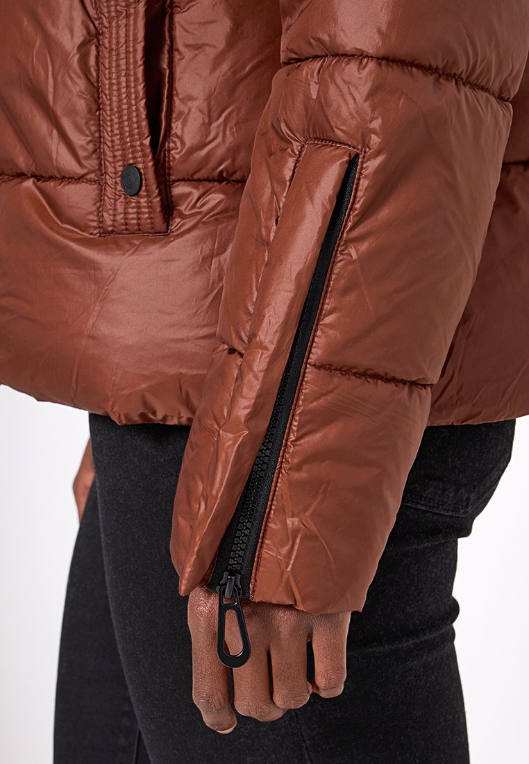 Giacca a vento marrone con finitura lucida, dettagli con zip sui polsini e cuciture ben visibili. Il tessuto sembra texturizzato e leggero.