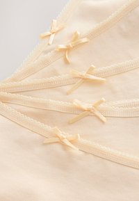 Pila de ropa interior de algodón beige con bordes ondulados y pequeños lazos de satén en cada cintura, exhibida sobre un fondo neutro.
