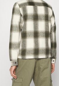 Camisa de franela en un patrón de cuadros en crema y verde olivo. Presenta un corte recto, puños con botones y un acabado de textura suave.