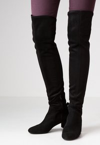 Botas de ante negras por encima de la rodilla con un diseño ajustado, un acabado texturizado y un tacón grueso. Cuenta con un zip lateral para ponerlas y quitarlas fácilmente.