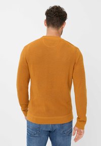 Senfgelbes Strickpullover mit langen Ärmeln, gerippter Textur, Rundhalsausschnitt und engen Bündchen, kombiniert mit blauen Jeans.