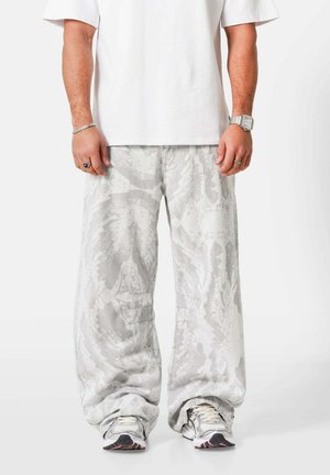 Man die losse, lichtgrijze broek met patroon draagt, witte sneakers en een effen wit T-shirt met korte mouwen, staand tegen een witte achtergrond.