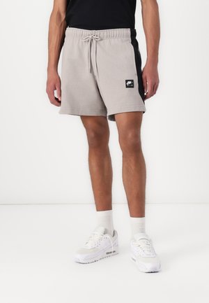 Grijze katoenen korte broek met een zwart zijpaneel, elastische tailleband en een logo patch aan de voorkant. Gecombineerd met witte sneakers en sokken tot mid-kuit.