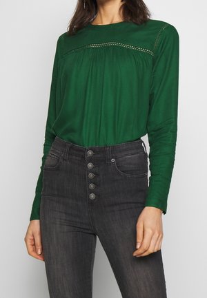 Frau trägt eine grüne Langarmbluse mit Spitzen-Detail und hoch taillierte dunkelgraue Jeans mit fünf vorderen Knöpfen.