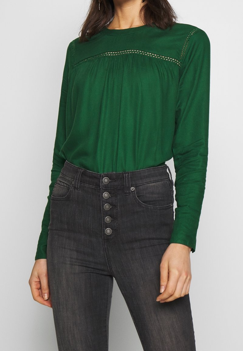 Femme portant un chemisier vert à manches longues avec un détail en dentelle et un jean gris foncé taille haute avec cinq boutons sur le devant.