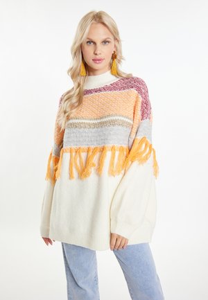 Blonde Frau trägt einen mehrfarbigen Strickpullover mit Fransen und gelben Quastenohrringen, kombiniert mit hellblauen Jeans, stehend vor weißem Hintergrund.