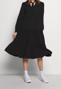 Robe noire à volants en tissu doux, dotée de manches longues, d'un col à nouer et d'un ourlet fluide. Associée à des baskets blanches et des chaussettes.