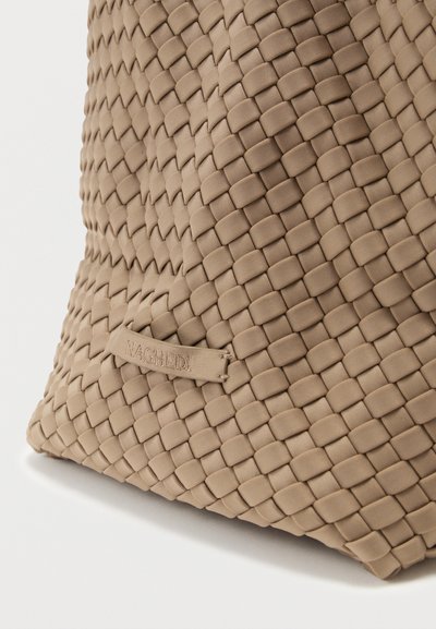 Μπεζ υφασμάτινη τσάντα με υφή σχεδίου και ετικέτα λογότυπου. Το υλικό φαίνεται μαλακό με περίπλοκο μοτίβο και χαλαρό σχήμα.
