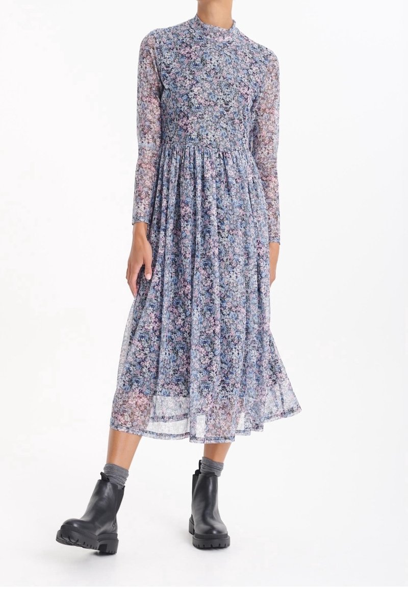 Femme portant une robe midi fluide à manches longues et col haut avec un motif floral bleu et rose, ainsi que des bottines noires avec des chaussettes grises.