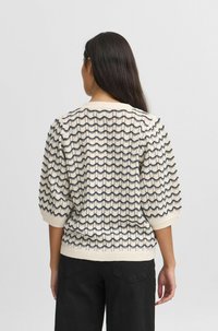 Pull crème avec motif ondulé bleu marine, manches bouffantes courtes, ourlet côtelé et tricot texturé. Vue de dos, porté sur un pantalon noir.