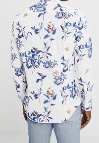 Biała koszula z długim rękawem, zdobiona niebiesko-pomarańczowym wzorem kwiatowym, z zaokrąglonym dołem i kołnierzykiem zapinanym na guziki.
