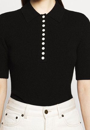 Frau trägt ein schwarzes kurzes Strick-Poloshirt mit Kragen und einer vertikalen Reihe weißer Knöpfe, kombiniert mit cremefarbenen Hosen.