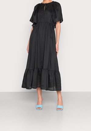 Femme portant une robe midi noire avec des manches courtes bouffantes et un ourlet volanté, associée à des sandales à talons ouvertes bleu clair.