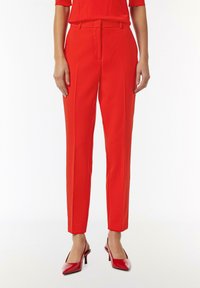 Red tailored trousers with a high waist, slim fit, and front pleats, paired with glossy red pointed-toe heels. Smooth fabric texture.