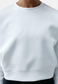 Sweatshirt blanc avec un col rond, en tissu texturé et avec des lettres en relief subtiles à l'avant. Doté d'un ourlet côtelé.