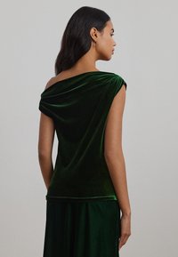 Grønn fløyelsbluse med en drapert off-the-shoulder design. Korte ermer og en jevn tekstur, kombinert med et matchende grønt skjørt.