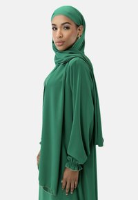 Grön hijab och långärmad klänning i lätt tyg. Har samlade ärmar och en flödande silhuett, med subtila guldfärger.