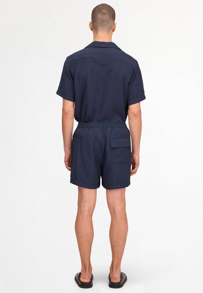 Man wearing navy short-sleeve shirt and matching elastic waist shorts with back pocket, standing on white background, facing away.