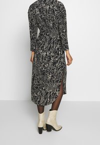 Abito lungo nero a maniche lunghe con un motivo floreale bianco, dotato di vita arricciata e spacchi laterali, abbinato a stivaletti beige texturizzati alla caviglia.