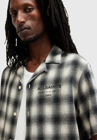 Chemise à carreaux noir et blanc avec un design à boutons et des manches courtes. Fabriquée dans un tissu texturé et comporte un logo bien visible sur la poitrine.
