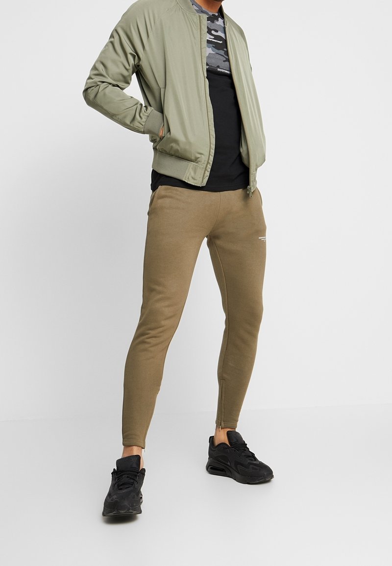 Olivgrüne Bomberjacke, schwarzes Camouflage-T-Shirt, tanfarbene Slim-Fit-Sweatpants, schwarze Sportschuhe, die ein lässiges Sportbekleidungsdesign hervorheben.