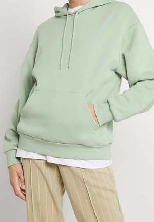 Persoon draagt een lichtgroene hoodie met een voorzak en beige pantalon met een smalle streep, met een wit shirt eronder.