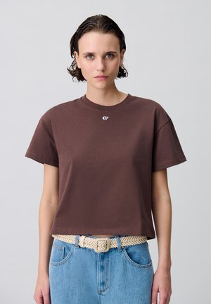 Camiseta corta marrón de algodón suave con mangas cortas y escote redondo, que presenta un pequeño logo blanco en el pecho. Combinada con jeans de mezclilla azules.