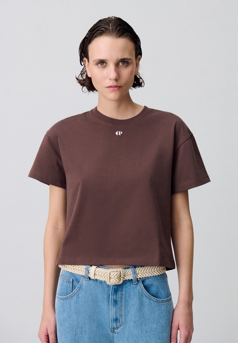 T-shirt cropped marron en coton doux avec des manches courtes et un col rond, présentant un petit logo blanc sur la poitrine. Porté avec un jean en denim bleu.