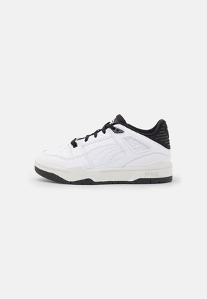 Biało-czarny niski sneaker z czarnymi sznurowadłami, wyściełanym kołnierzem i wzorzystą podeszwą na jednolitym jasnoszarym tle.